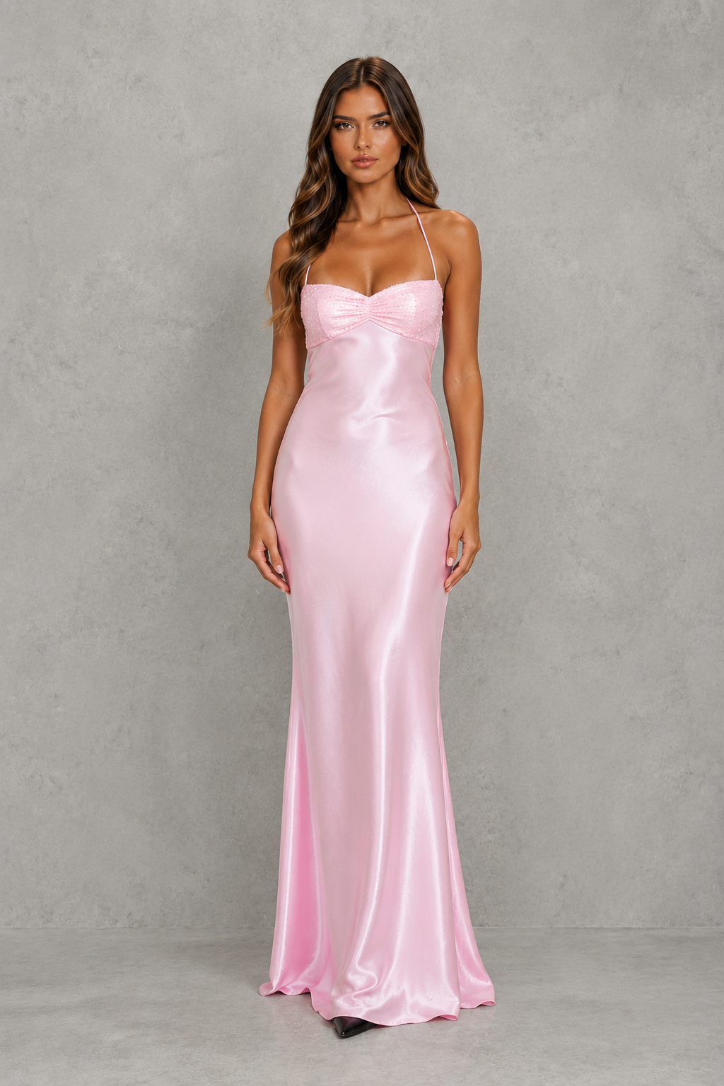 A woman modeling a floor-length light pink satin gown with spaghetti straps and a sweetheart neckline, standing against a textured gray studio backdrop.