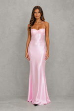 A woman modeling a floor-length light pink satin gown with spaghetti straps and a sweetheart neckline, standing against a textured gray studio backdrop.