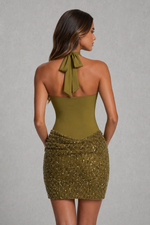A woman shown from the back wearing a sleeveless olive green halter mini dress with a sequined body and a bow detail at the back of the neck, set against a neutral gray background.
