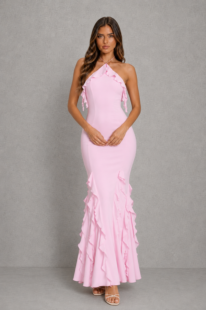 A woman modeling a light pink halter-neck maxi dress with ruffle details along the neckline and cascading down the skirt, set against a textured gray studio background.
