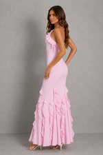 A woman modeling a light pink sleeveless maxi dress with ruffle details, standing against a neutral gray studio background.