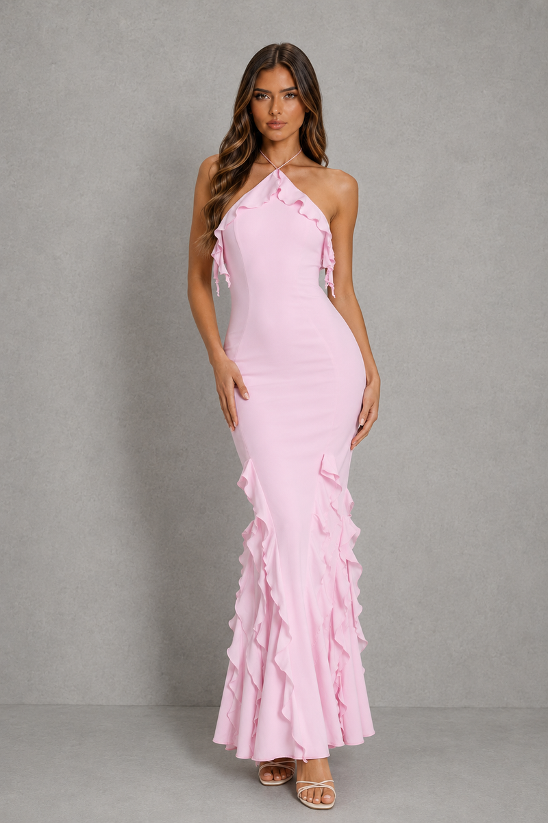 A woman modeling a light pink halter-neck maxi dress with ruffle details along the neckline and flared hem, standing against a textured gray studio backdrop.