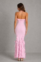 A woman viewed from the back wearing a light pink strapless halter maxi dress with ruffle trim at the neckline and along the flared hem, photographed against a plain gray studio backdrop.