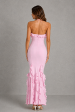 A woman viewed from the back wearing a light pink strapless halter maxi dress with ruffle trim at the neckline and along the flared hem, photographed against a plain gray studio backdrop.