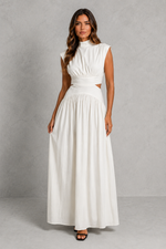 A woman modeling a sleeveless white maxi dress with a high mock neck, gathered waist, and side cutouts, standing against a textured gray studio backdrop.
