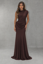A woman modeling a floor-length, high-neck draped gown in a deep chocolate brown color, standing against a textured gray studio backdrop.