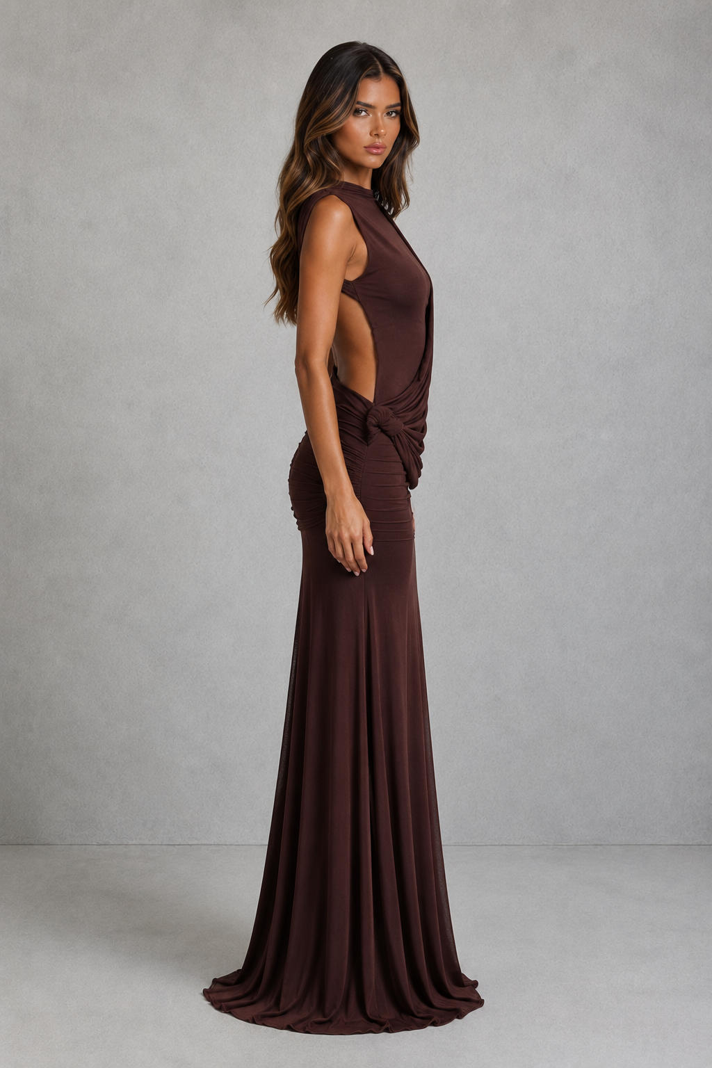 A woman modeling a floor-length, sleeveless dark brown gown with a high neckline, side cutout, and draped waist detail, photographed against a neutral gray studio backdrop.