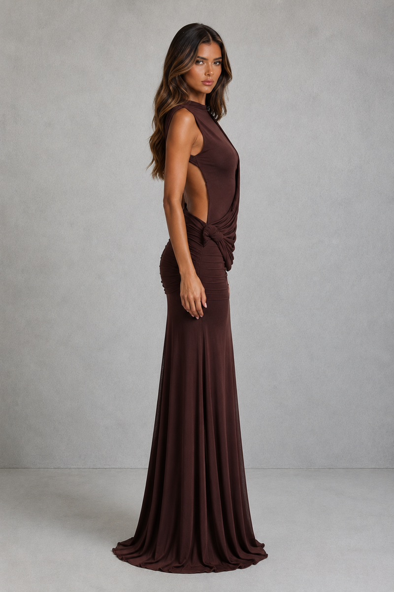 A woman modeling a floor-length, sleeveless dark brown gown with a high neckline, side cutout, and draped waist detail, photographed against a neutral gray studio backdrop.