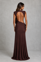 A woman modeling a floor-length, sleeveless gown from the back, showing a draped open back and gathered skirt in a dark brown shade, set against a textured gray studio backdrop.