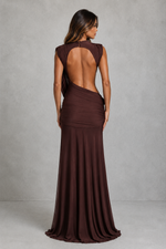 A woman modeling a floor-length, sleeveless gown from the back, showing a draped open back and gathered skirt in a dark brown shade, set against a textured gray studio backdrop.