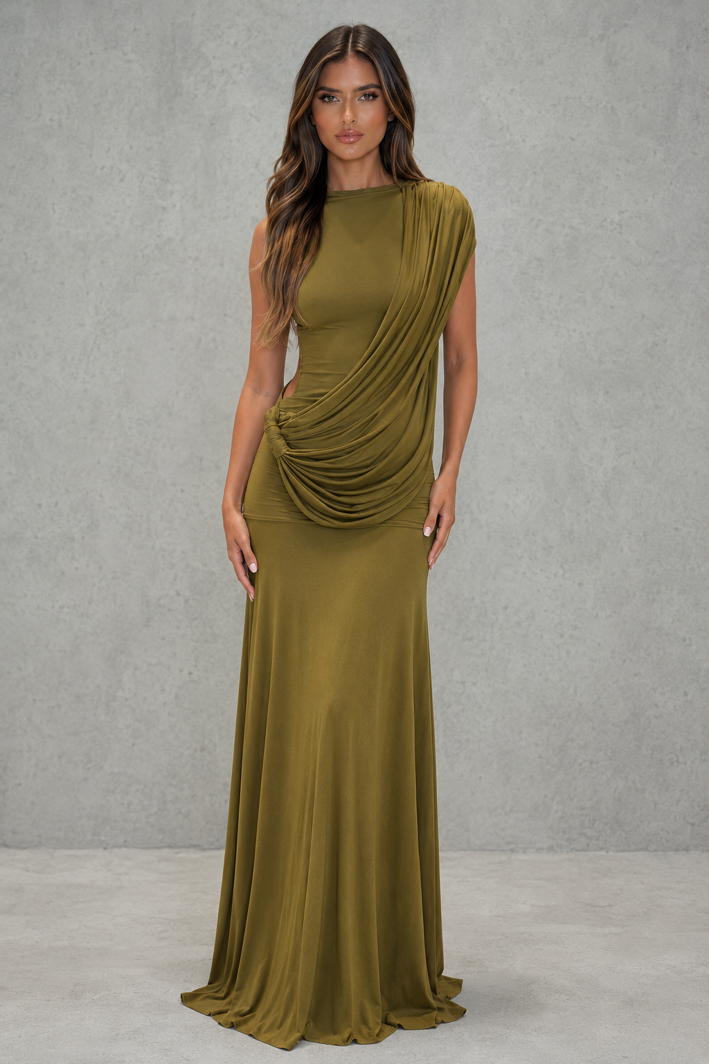 A woman modeling a floor-length draped gown in an olive green color, standing against a textured gray studio wall.