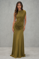 A woman modeling a floor-length draped gown in an olive green color, standing against a textured gray studio wall.