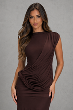 A woman modeling a sleeveless, draped brown dress against a textured gray studio background.