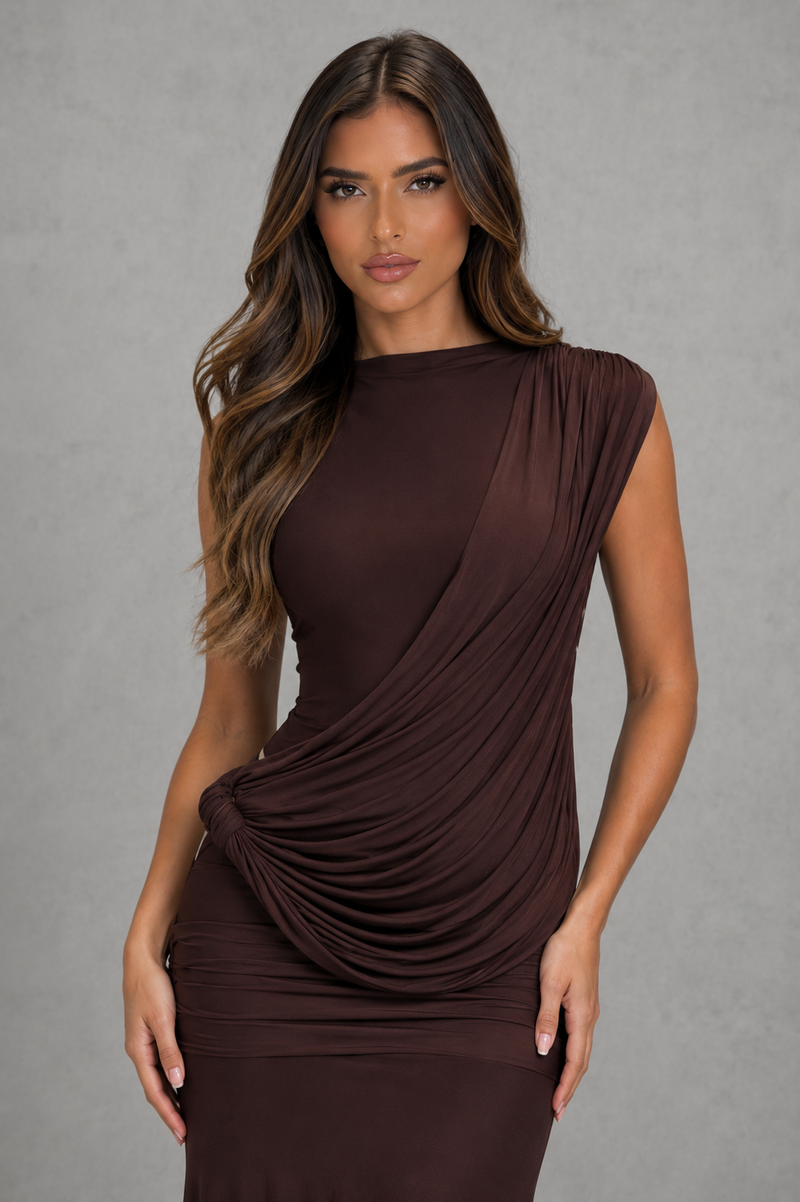 A woman modeling a sleeveless, draped brown dress against a textured gray studio background.