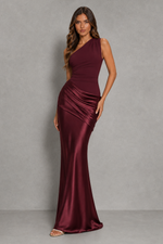 A woman modeling a floor-length, one-shoulder satin gown in deep burgundy against a textured gray studio backdrop.