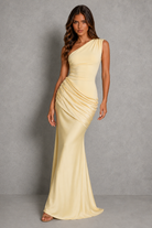 A woman modeling a floor-length, one-shoulder satin gown in a pale pastel yellow against a textured gray studio backdrop.