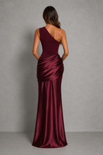 A woman viewed from the back wearing a floor-length satin gown in deep burgundy. The dress has an asymmetrical neckline and a draped ruched waist, set against a neutral gray studio background.