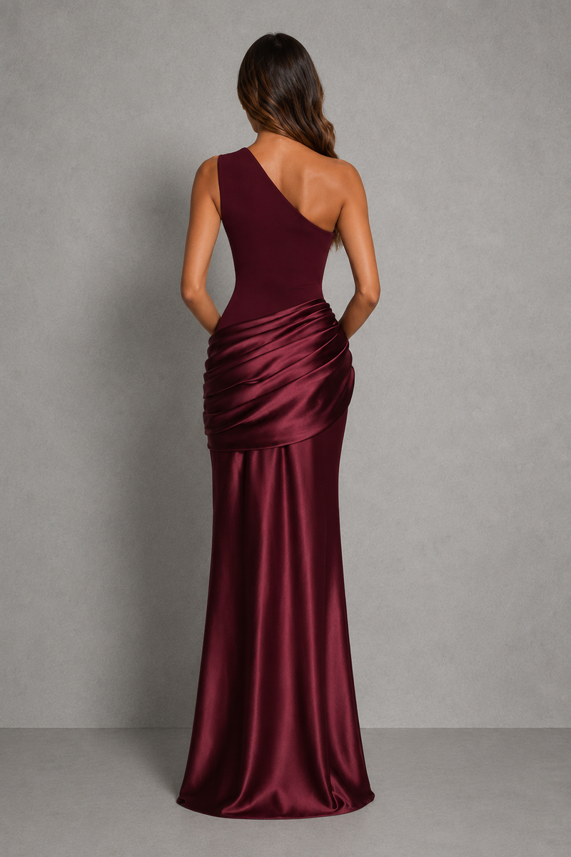 A woman viewed from the back wearing a floor-length satin gown in deep burgundy. The dress has an asymmetrical neckline and a draped ruched waist, set against a neutral gray studio background.