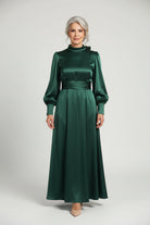 Person wearing a long green dress standing in a room with a sofa and white curtain.