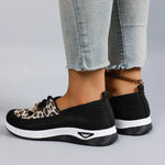 Black slip-on sneakers with leopard print accents worn with light blue jeans on a gray background