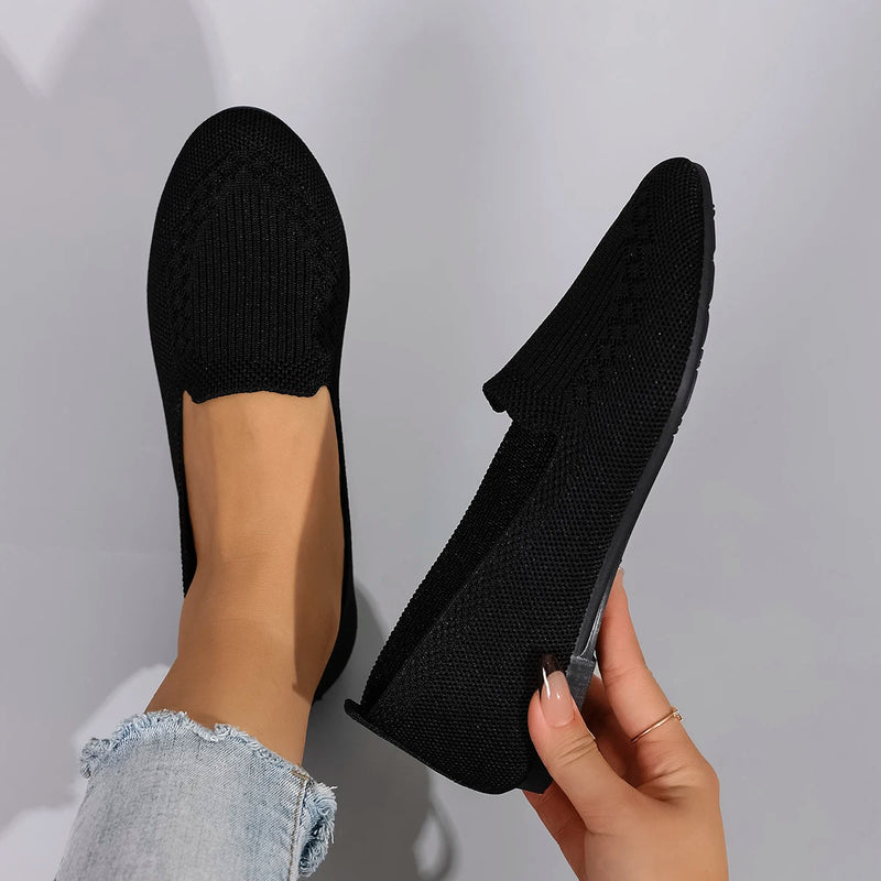Black slip-on shoes being held by a hand against a light gray background