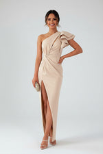 Woman wearing a beige one-shoulder dress with a slit, standing in a room with a white wall and beige sofa.