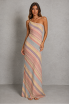 A woman modeling a one-shoulder maxi dress with diagonal pastel stripes in pink, beige, and light blue-gray against a textured gray studio backdrop.