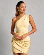 A woman modeling a pale yellow one-shoulder ruched mini dress against a textured gray studio background.