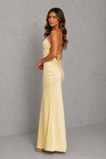 A woman modeling a pale yellow satin maxi dress with a backless design, thin shoulder straps, and side tie details, set against a textured gray studio background.