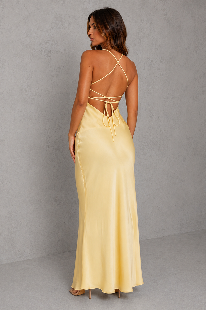 A woman modeling a pale yellow satin maxi dress from the back, featuring a low open back with crisscrossing tie straps, set against a textured gray studio background.