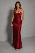 A woman modeling a floor-length satin slip dress in deep burgundy red, standing against a textured gray studio backdrop.