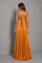 Women’s golden orange pleated halterneck maxi dress with a cross-front neckline, open back, and flowing floor-length silhouette for wedding guests and formal occasions