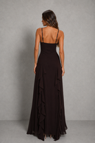 A woman wearing a long black spaghetti-strap maxi dress with a ruffled hem, shown from the back against a textured gray studio backdrop.