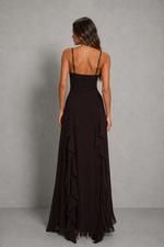 A woman wearing a long black spaghetti-strap maxi dress with a ruffled hem, shown from the back against a textured gray studio backdrop.