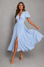 A woman modeling a light blue midi dress with a deep V-neckline, short puff sleeves, a gathered waist, and a button-front skirt with a thigh-high slit, posed against a plain gray studio backdrop.