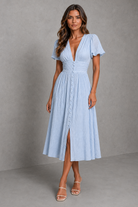 A woman modeling a light blue midi dress with a smocked bodice, short puff sleeves, a deep V-neckline, and a button-down front, set against a textured gray studio backdrop.