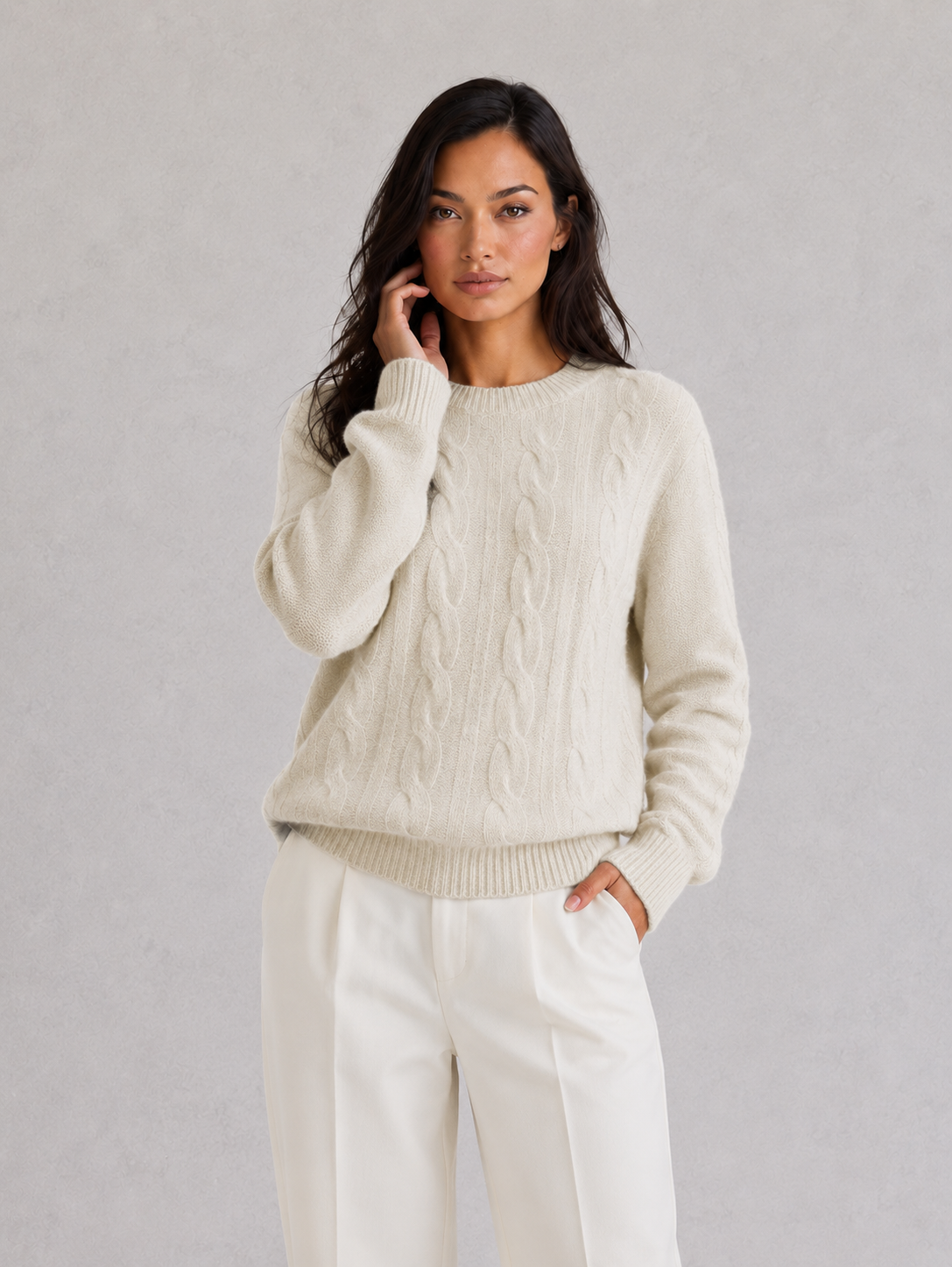 Woman wearing a cream sweater and white pants against a gray background