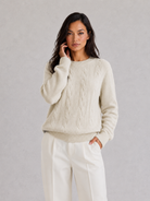 Woman wearing a cream sweater and white pants against a gray background