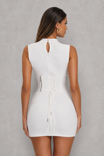 A woman shown from the back wearing a fitted sleeveless white mini dress with a high neckline, keyhole cutout at the back, and lace-up detailing at the waist, against a textured gray wall.