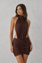 A woman modeling a dark brown halter-neck mini dress with side cutouts and a ruched wrap-style skirt, posed against a textured light gray studio background.