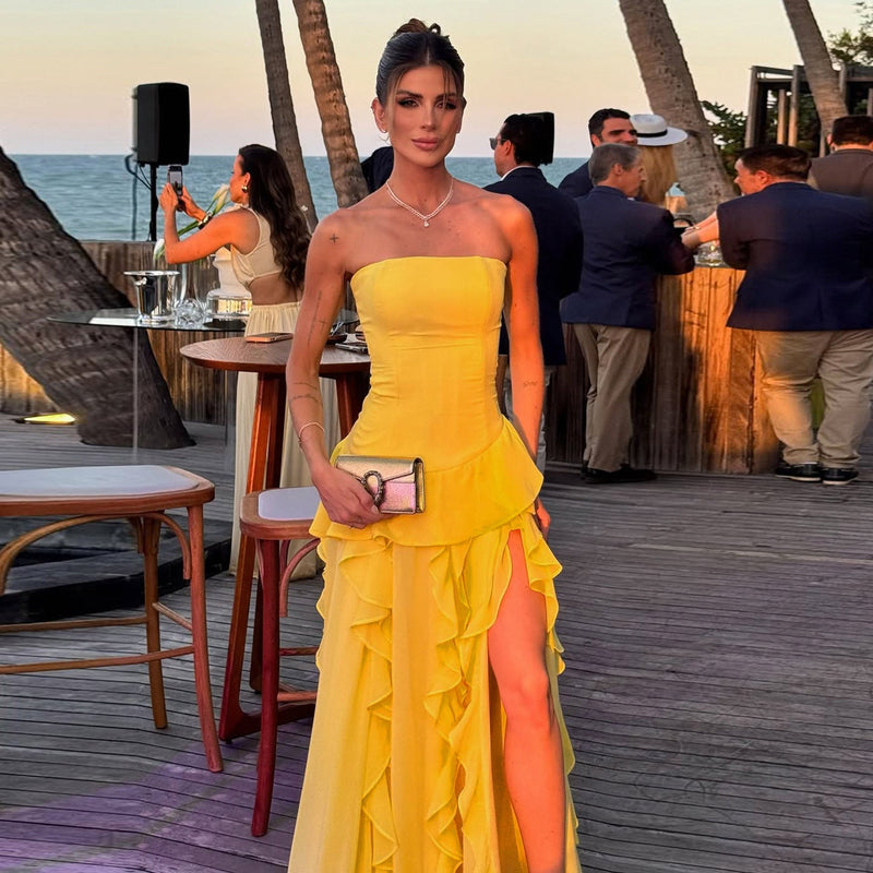 Woman in a yellow strapless dress with an orange slit standing on a wooden deck by the sea.
