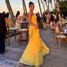 Woman in a yellow dress standing on a wooden deck with palm trees and people in the background.