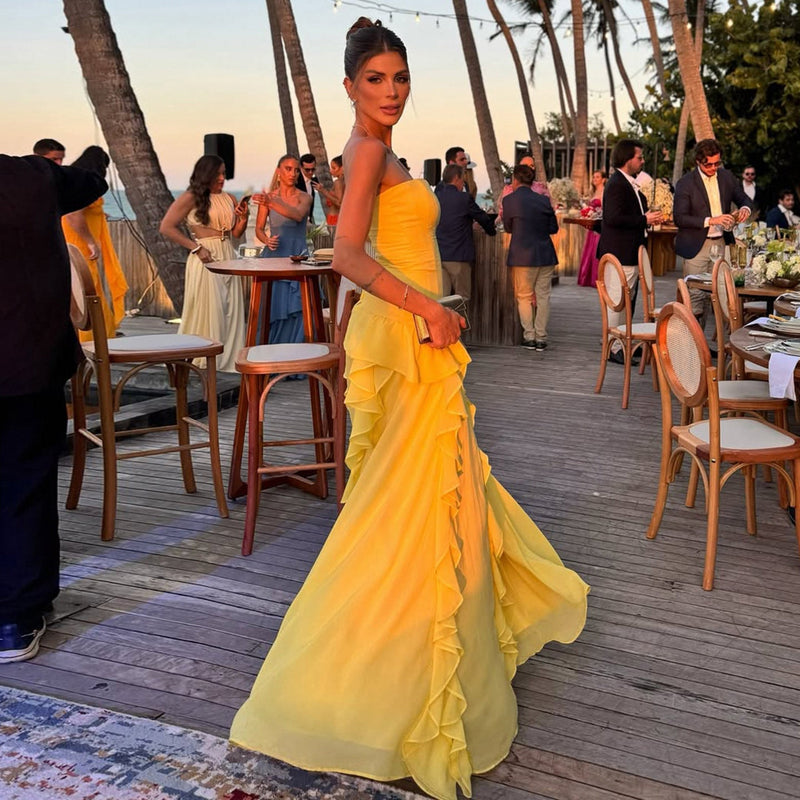 Woman in a yellow dress standing on a wooden deck with palm trees and people in the background.
