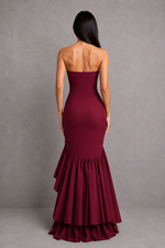Woman wearing a burgundy strapless gown with ruffled skirt against a gray background