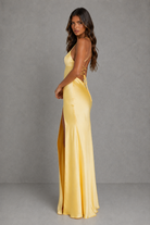 A woman modeling a floor-length yellow satin gown with a high side slit and lace-up back detail, photographed against a plain gray studio backdrop.