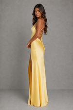A woman modeling a floor-length yellow satin gown with a high side slit and lace-up back detail, photographed against a plain gray studio backdrop.