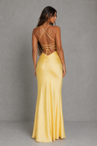 A woman modeling a floor-length yellow satin gown from the back, showing a strappy open back and flared skirt, against a gray studio backdrop.