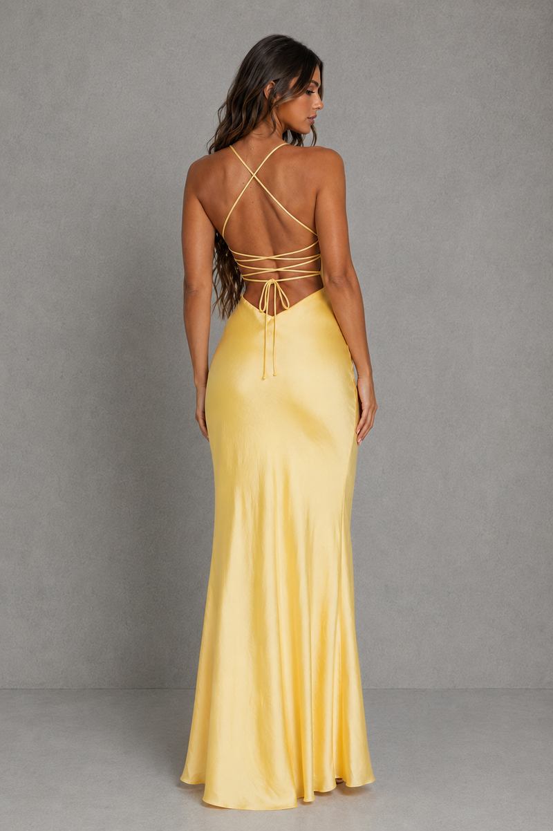 A woman modeling a floor-length yellow satin gown from the back, showing a strappy open back and flared skirt, against a gray studio backdrop.