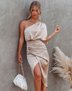 Woman in a beige one-shoulder dress holding a white handbag against a gray background