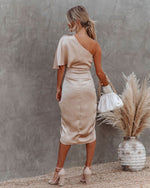 Woman in a beige one-shoulder dress standing against a gray wall with dried pampas grass and a vase.